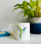 White mug with a colorful bird design on a surface with a plant in the background